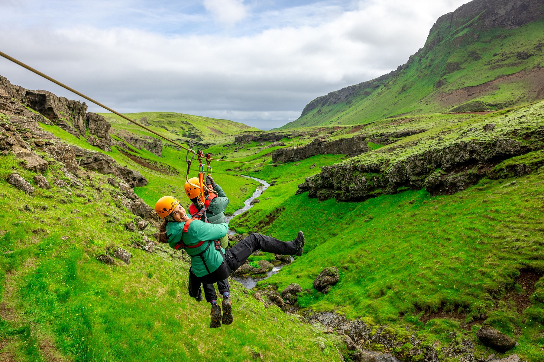 Two women ziplining over an open grassy field, with mountains in the distance