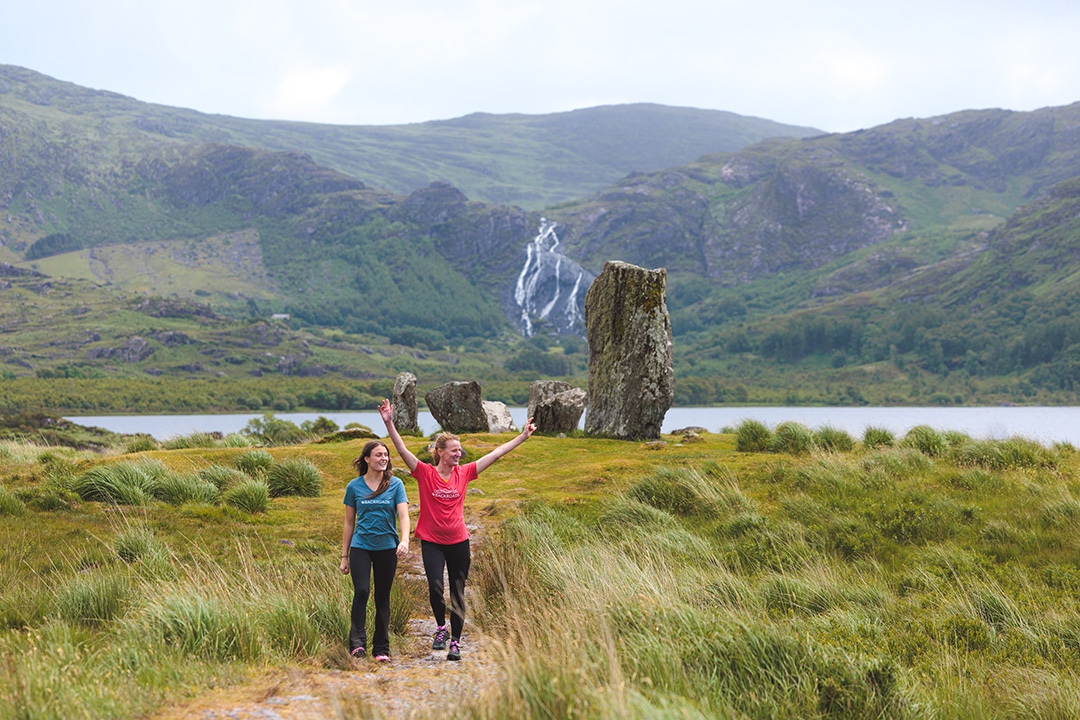Two women in a field of tall grass with their arms open, with tall boulders in the background