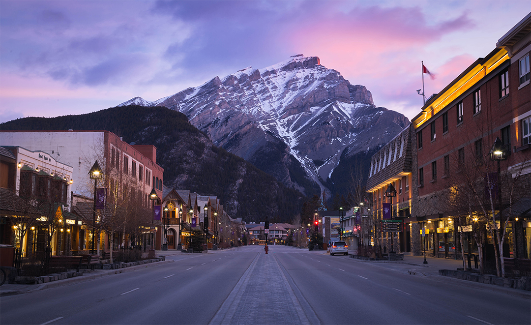 Town with empty roads and shop buildings to the left and right, with snow-capped mountains in the distance