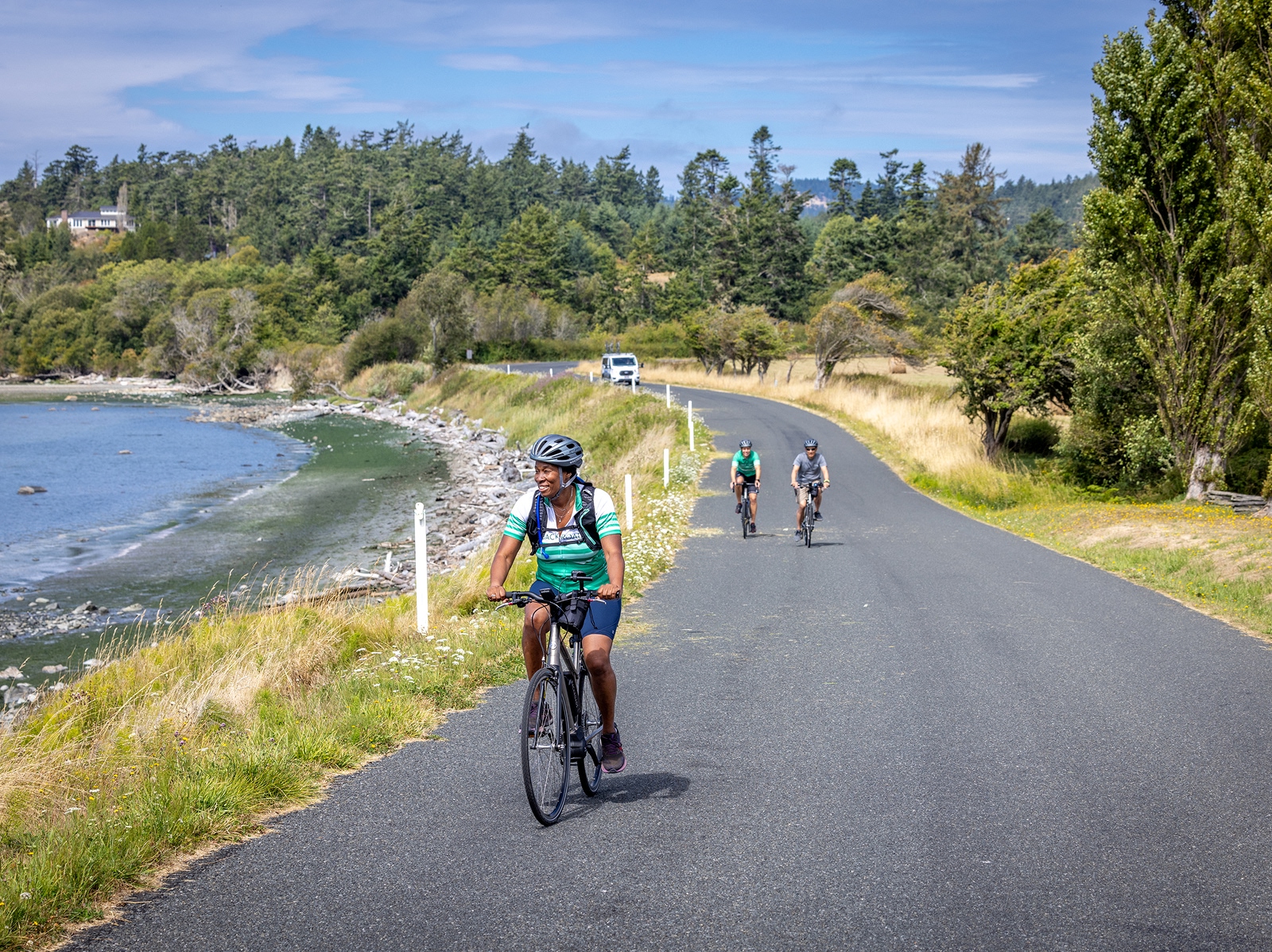 Three people biking on an empty road, with the ocean to the left and large trees in the background