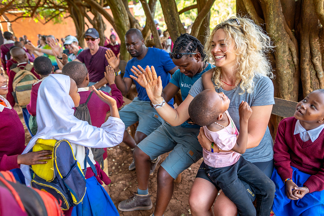Woman holding a baby, giving high-fives to a group of children walking by