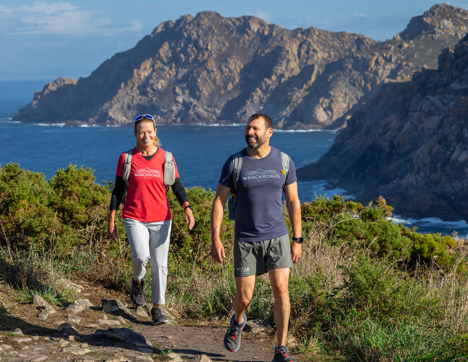 Man and woman smiling while walking on a dirt and stone path and the ocean in the background