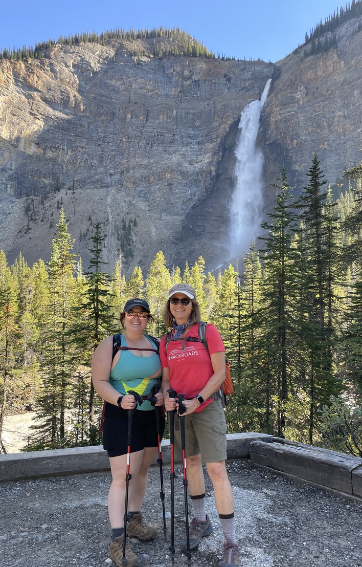 Two women smiling while holding hiking poles in front of a large forest