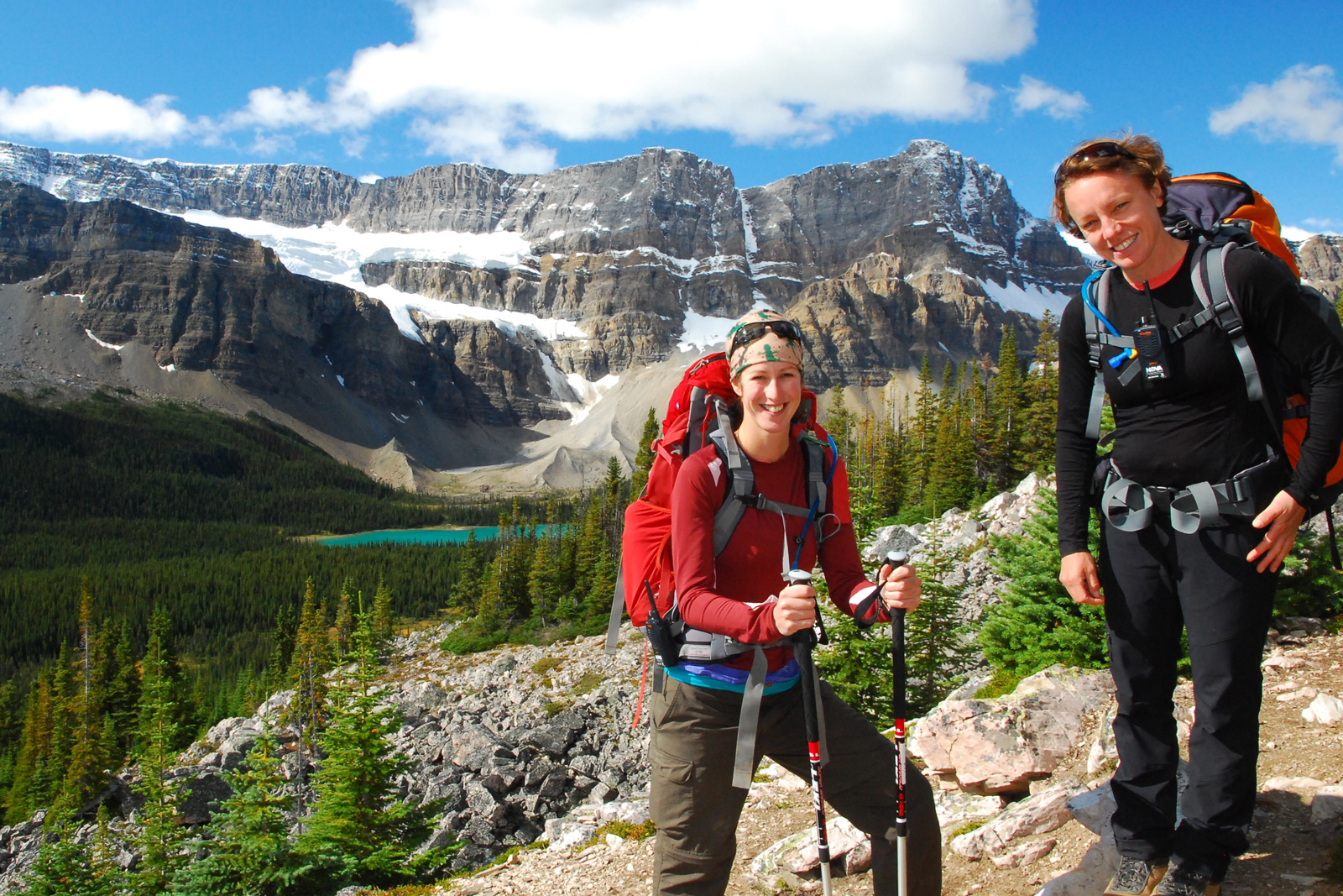 Two women with large hiking backpacks, standing on large boulders in a valley