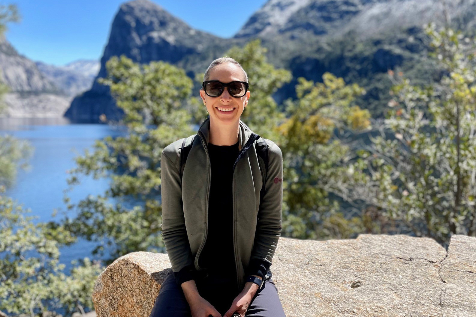 Woman smiling and wearing sunglasses, sitting on a boulder with a lake in the distance