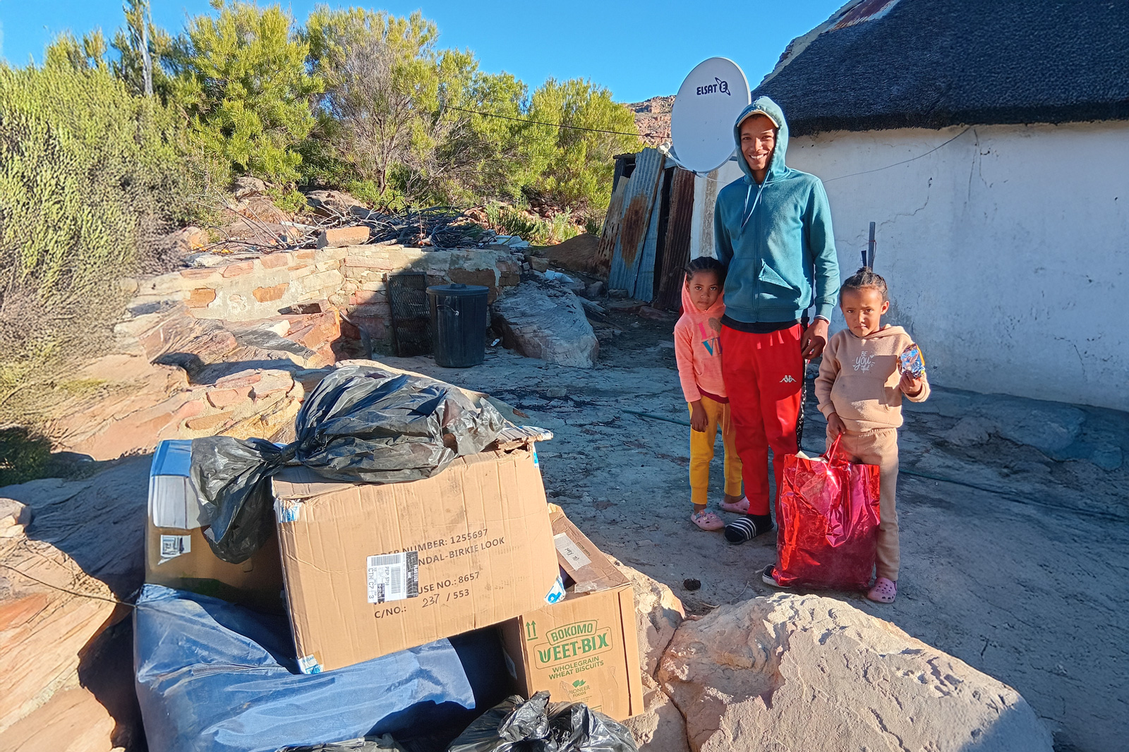 Man and child standing next to large piles of plants and boxes in a desert