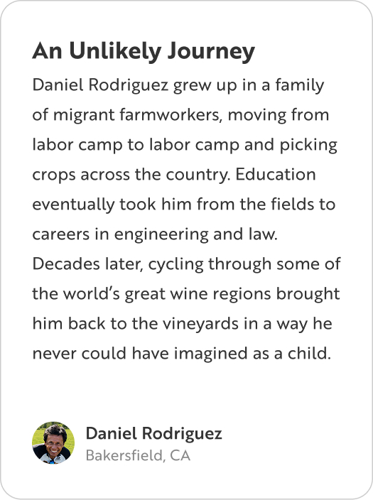 A profile card titled "An Unlikely Journey" from Daniel Rodriguez of Bakersfield, CA. The text reads: "Daniel Rodriguez grew up in a family of migrant farmworkers, moving from labor camp to labor camp and picking crops across the country. Education eventually took him from the fields to careers in engineering and law. Decades later, cycling through some of the world's great wine regions brought him back to the vineyards in a way he never could have imagined as a child."