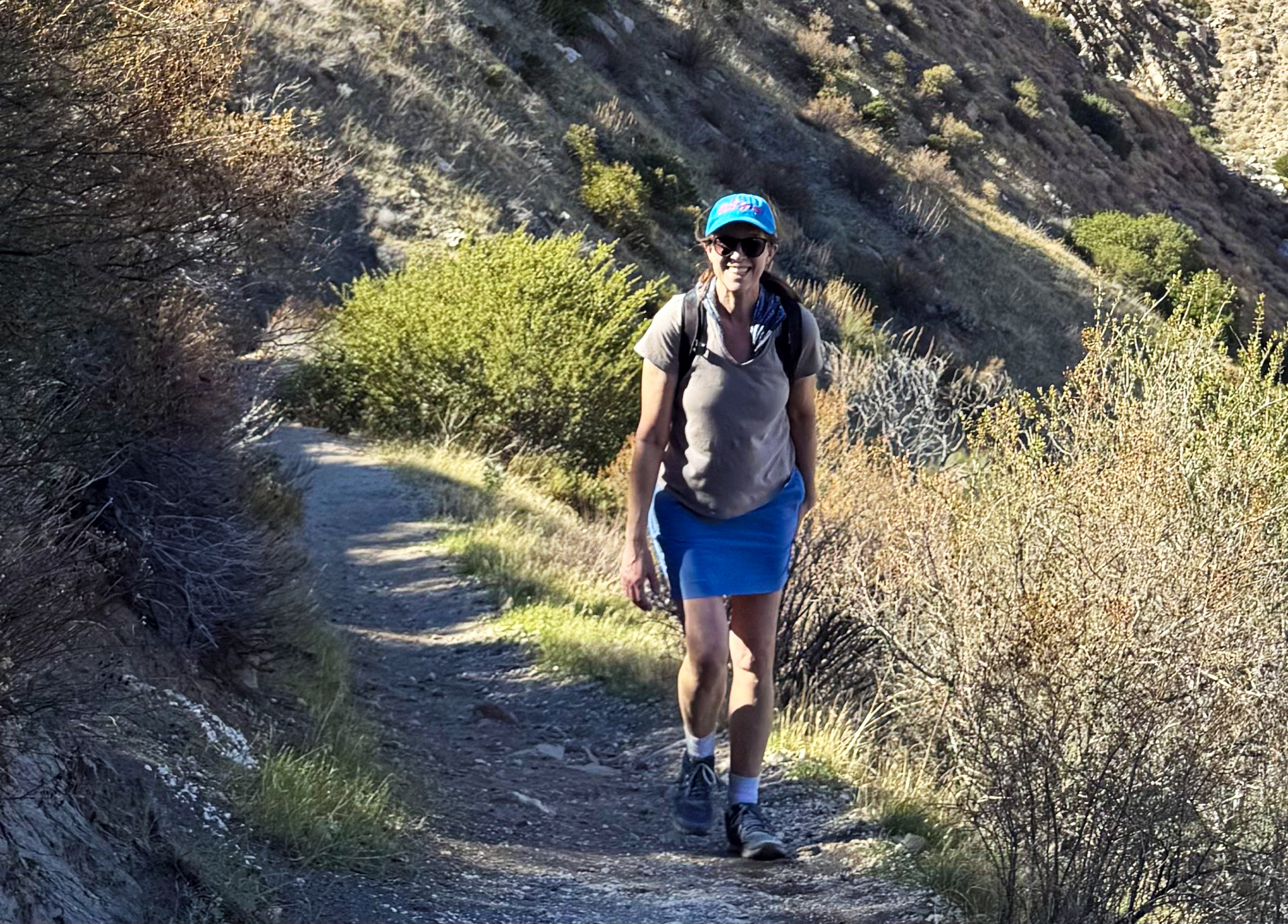 Woman wearing a blue hat while smiling, hiking up a gravel trail