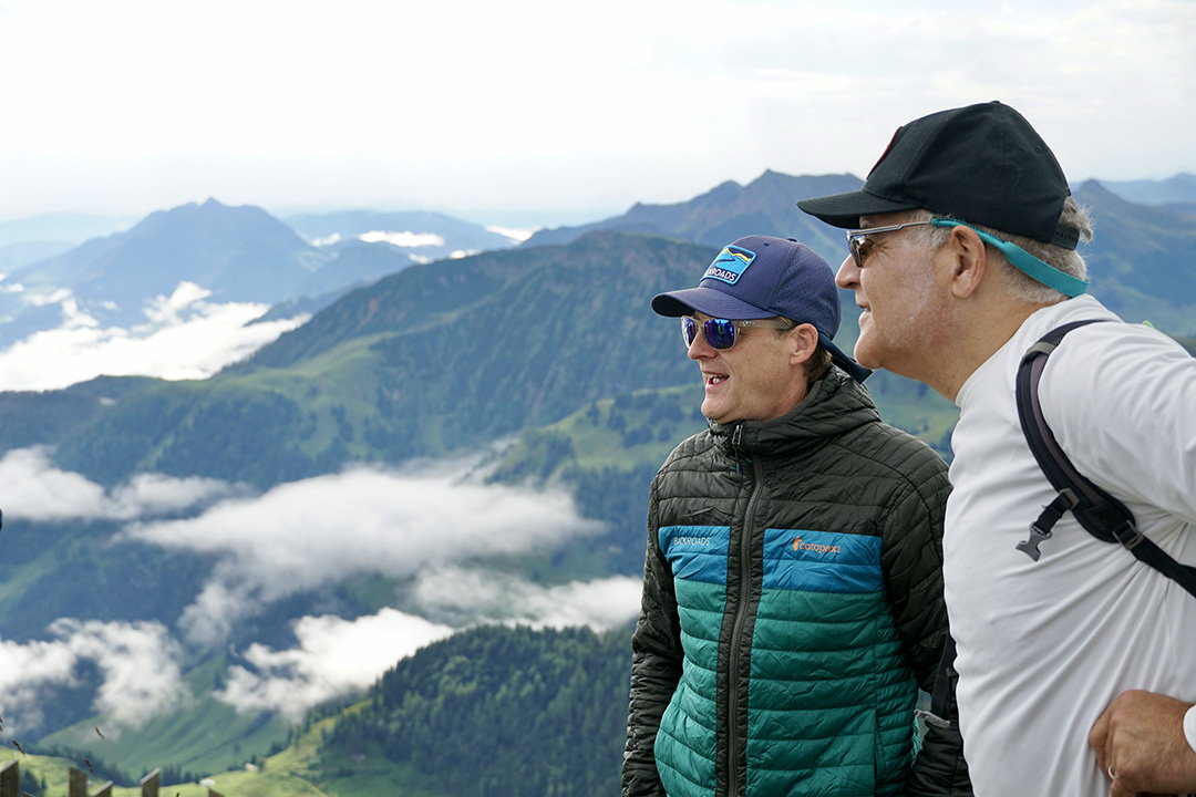 Two men wearing sunglasses on top of a mountain, looking towards a large, foggy valley