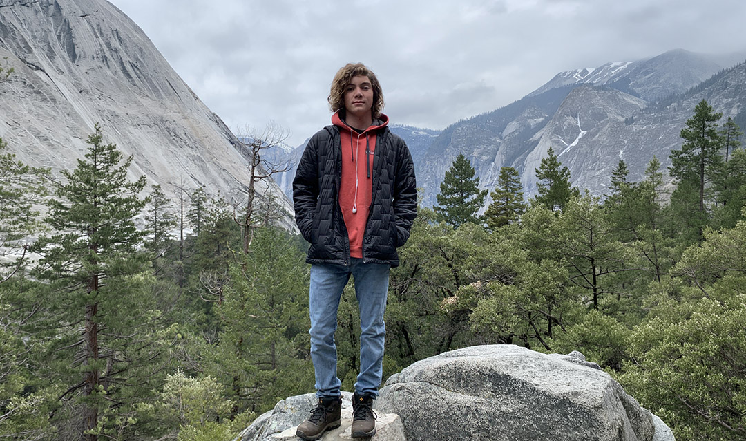 Man with a salmon colored sweater, standing on top of a tall boulder with a forest in the background