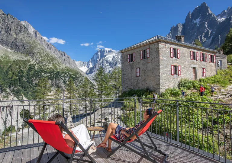 Two women sitting on reclining chairs on a wooden balcony, looking out to large mountains