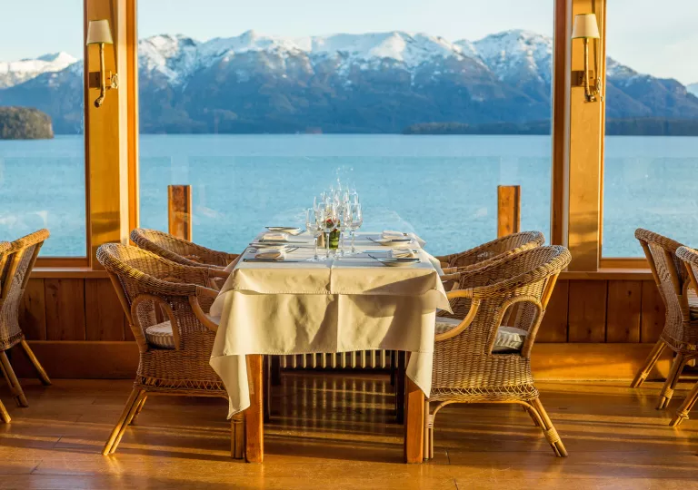 Outdoor dining table with woven chairs, a white table looking out towards a large windows to tall mountains