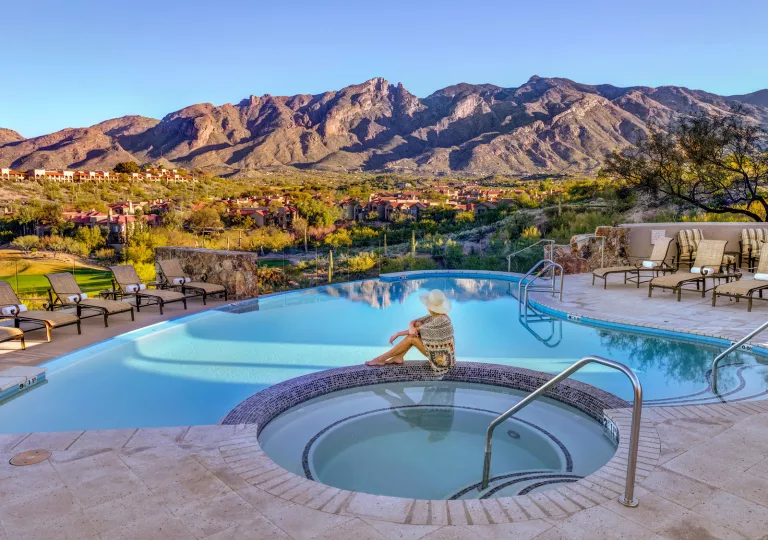 Woman sitting on stone in between a pool and hut tub, looking out towards tall mountains