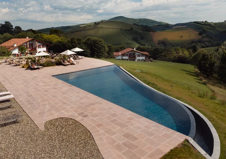 Outdoor pool with brick walkway to the left and pool chairs by a building