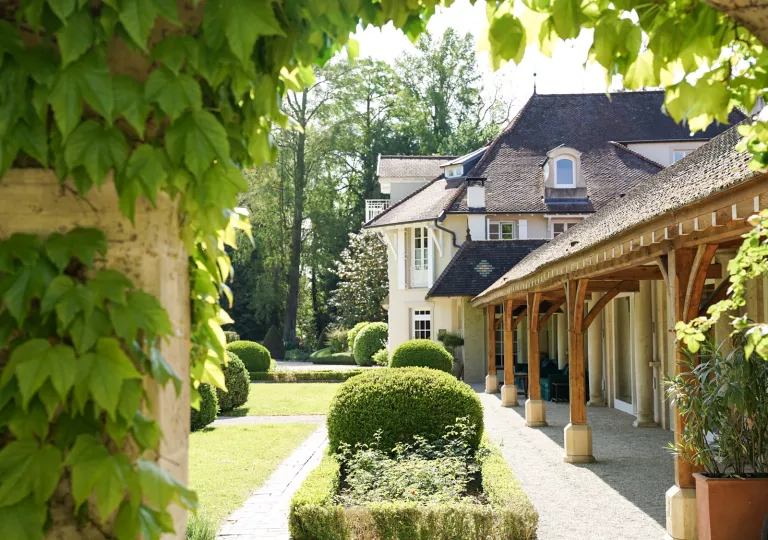 Outdoor view of white and brown building with bushes and tall trees in the background