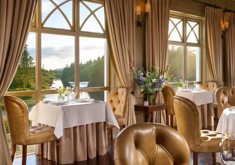 Dining area with cushioned chairs and table cloths over square tables, looking out towards a lake with tall trees
