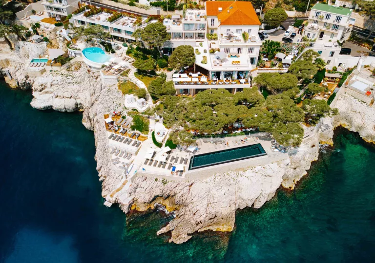 Sky view of a hotel complex with an outdoor pool on a cliff, next to the ocean