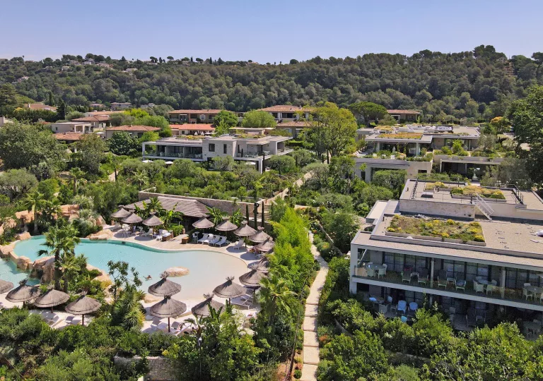 Outdoor pool surrounded by straw umbrellas, next to large hotel villa buildings