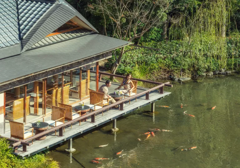 Man and woman sitting on a wooden balcony, looking out to a pond filled with koi fish
