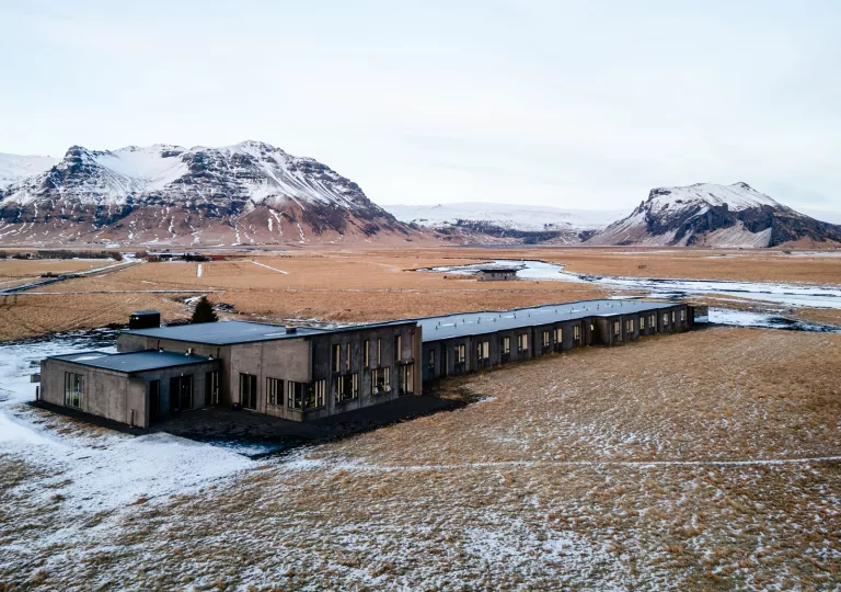 Exterior view of gray building in the middle of a valley, with snowy mountains in the background