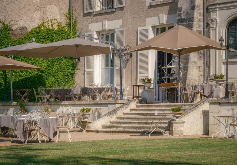 Outdoor patio area with white metal chais, brown umbrellas and a stone building on the right