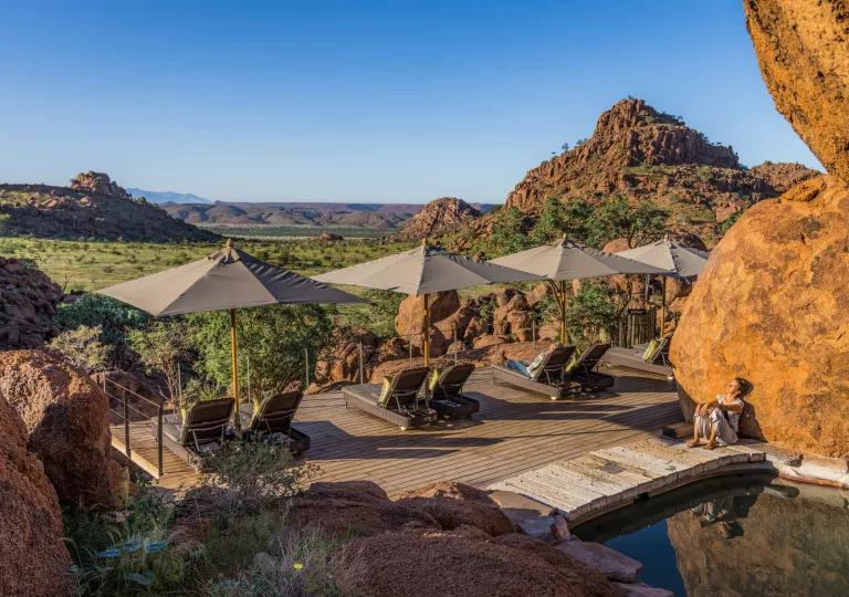 Reclining chairs with umbrellas on a wooden patio, overlooking a large, grass valley with boulders