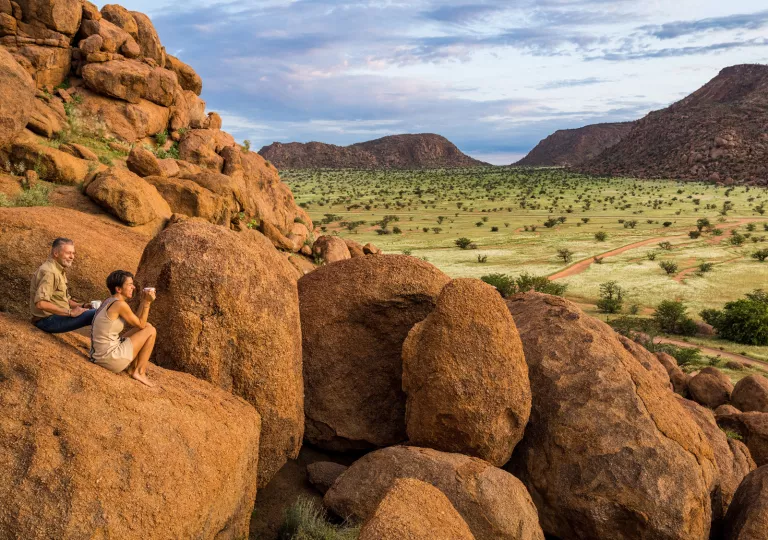 Man and woman sitting on a large boulder overlooking a large, grassy valley