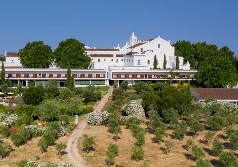 Exterior view of wide, white building with red balconies with a large valley of trees in front