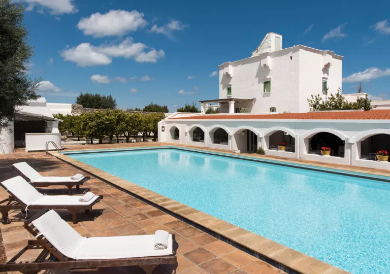 Outdoor pool, surrounded by white pool chairs and a white, rectangular building