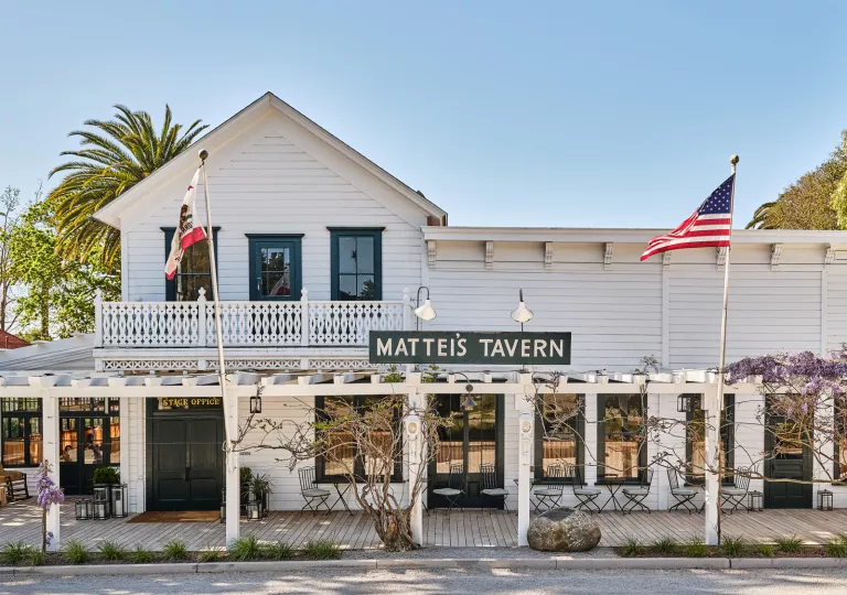 White building with a wooden porch and two flas on metal rods in front