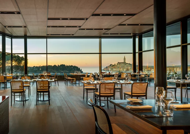 A hotel dining room with many tables and large windows overlooking the water