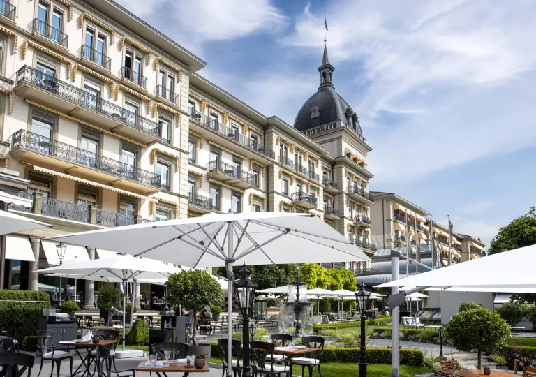 Tables and chairs under large umbrellas, next to a wide hotel building