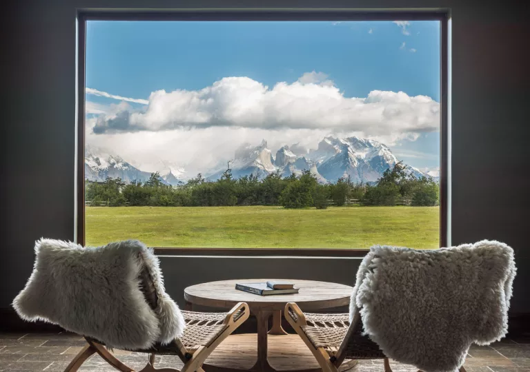 Two woven chairs with fur on them, facing a window looking out to snow capped mountains