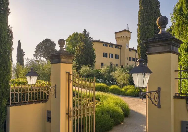 Exterior view of yellow and brown hotel building, with a golden fence in front of a garden