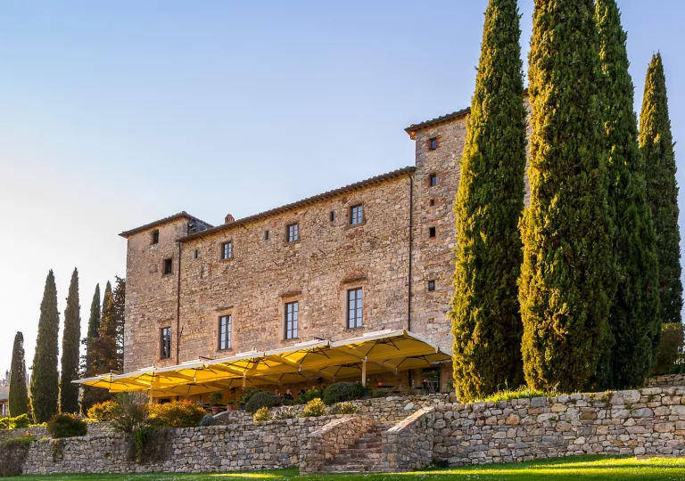 Tall stone building with tall trees and an outdoor patio covered by yellow umbrellas