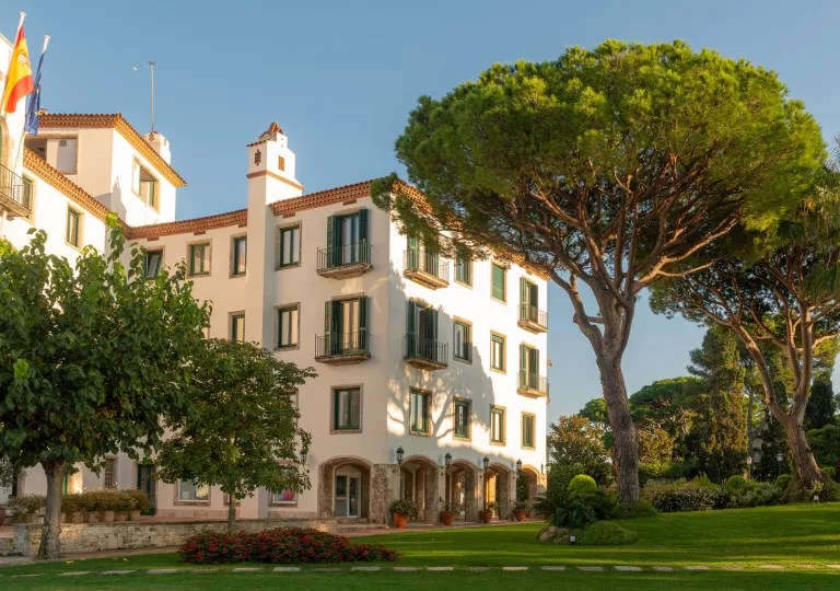 Exterior view of white and stone hotel building, surrounded by tall trees and a grass field