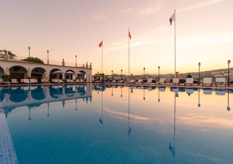 Wide, outdoor pool surrounded by white umbrellas and chairs, with the sunset in the background
