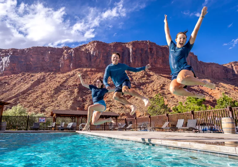 Three people jumping into an outdoor pool, with a canyon in the background