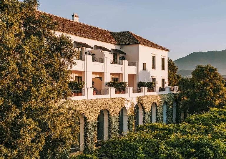 Exterior view of white and brown building, covered in plants and surrounded by trees