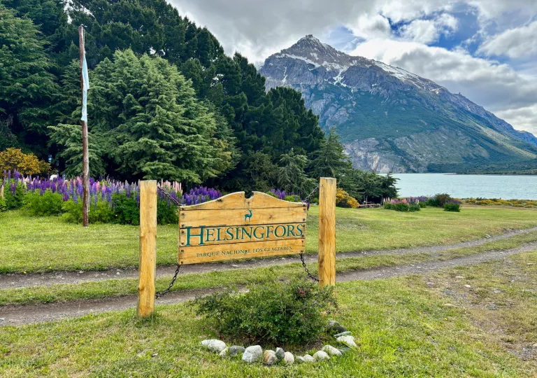 Wooden sign with the word "Helsingfors", in front of tall trees and an open lake