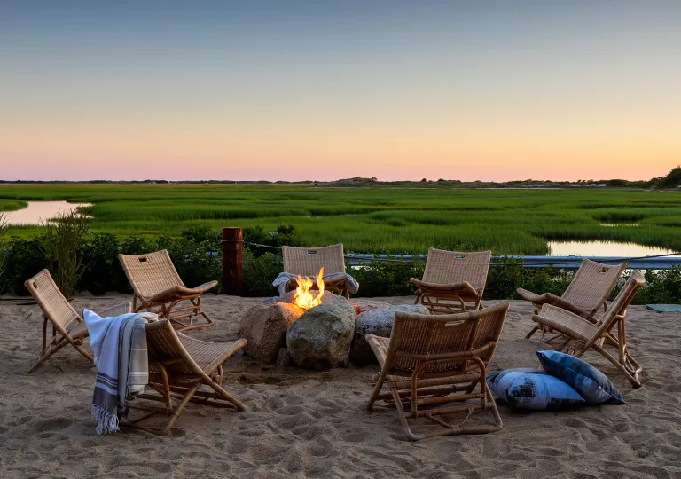 Sandy patio with woven chairs and a firepit, with a large marsh in the background