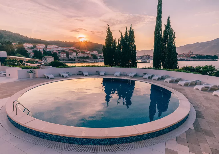 Outdoor, circular pool surrounded by reclining white chairs and a sunset in the background