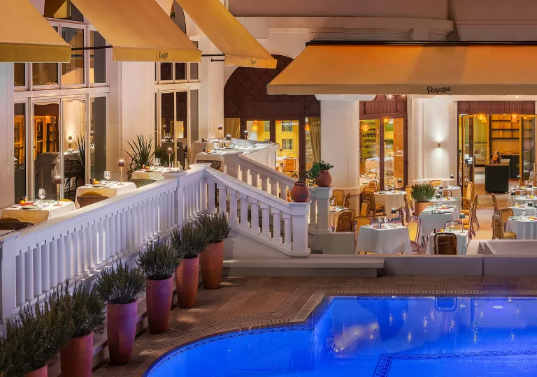 Outdoor patio of a hotel building with dining tables aside an illuminated pool