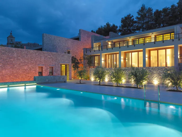 Night time view of an outdoor pool, with an illuminated building with large, glass walls and mini balconies