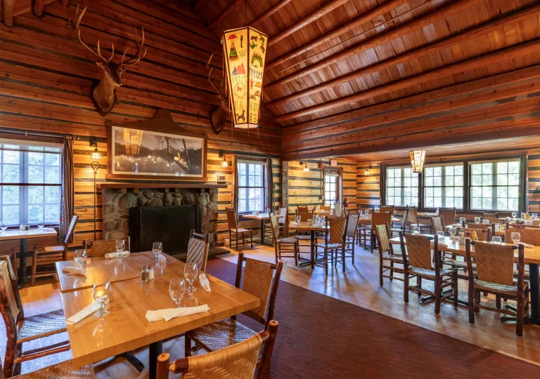 Lodge-style restaurant dining hall with wooden tables and chairs, with a deer head hanging over a fireplace
