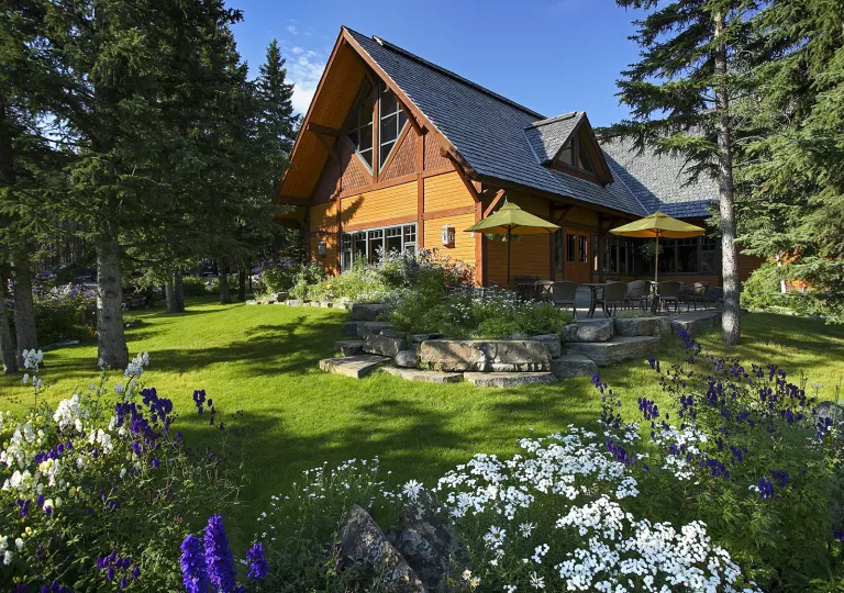 Exterior view of a wooden cabin with a garden and tall trees in front