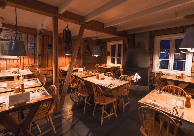 Dimly lit cabin restaurant with dining tables and wooden chairs, with a fireplace in the back