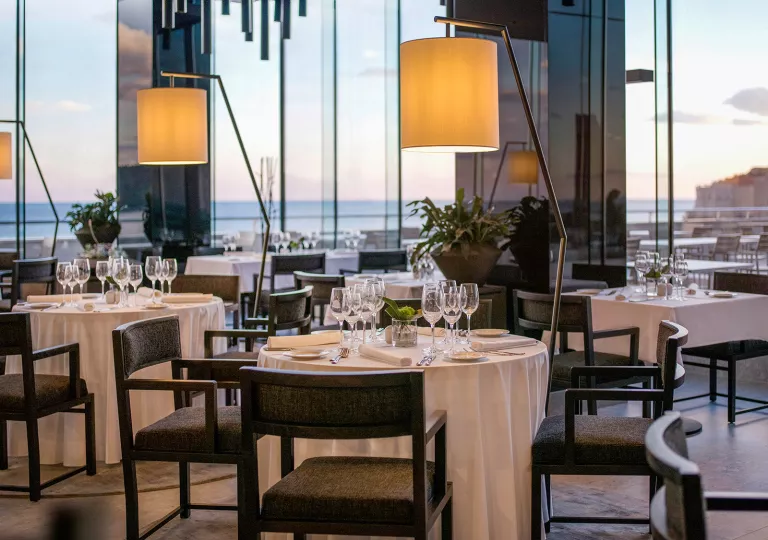 Restaurant dining hall with large glass walls, light fixtures over each table and wine glasses on each table