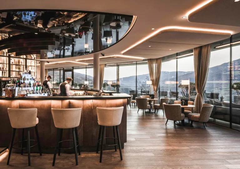 Indoor bar and lounge with two bartenders behind the counter, with beige stools and wooden tables in the room