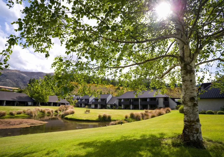 Exterior view of hotel buildings with large balconies, with a grass field and small lake in front